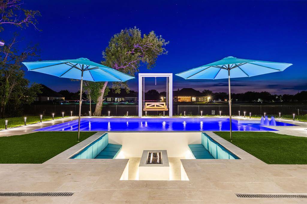 Experience luxury outdoor living with this breathtaking poolscape, featuring a glowing resort-style pool with water features and ambient lighting. A sunken fire pit lounge offers a cozy spot for evening gatherings, while oversized umbrellas provide shade and comfort. The modern swing and expansive backyard views complete this stunning space, perfect for entertaining or unwinding under the stars.