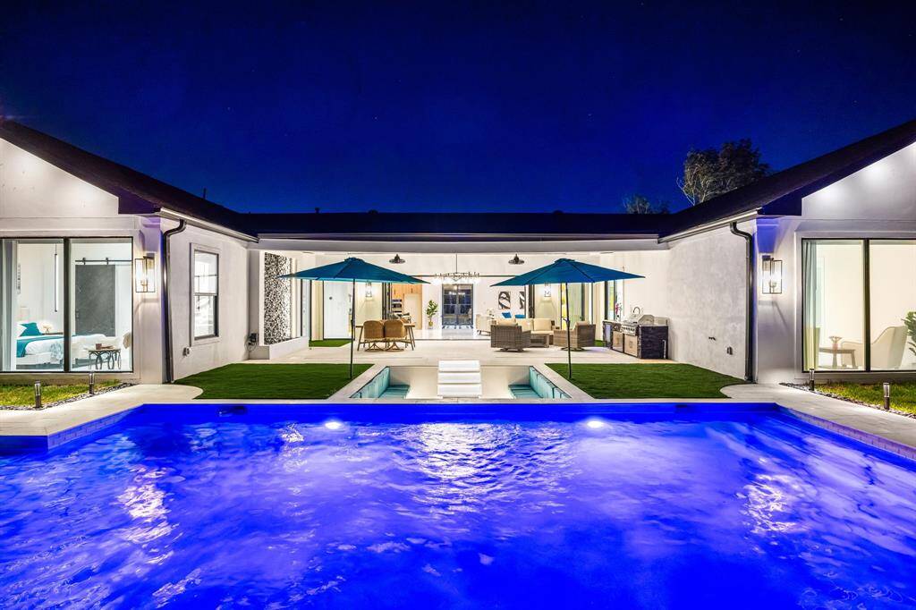 A stunning view of this contemporary home's backyard oasis showcases a beautifully illuminated pool leading up to a covered patio with built-in lighting and ceiling fans. The outdoor area includes a sunken fire pit lounge, lush grass, and a fully equipped outdoor kitchen—perfectly designed for seamless indoor-outdoor living and upscale entertaining under the stars.