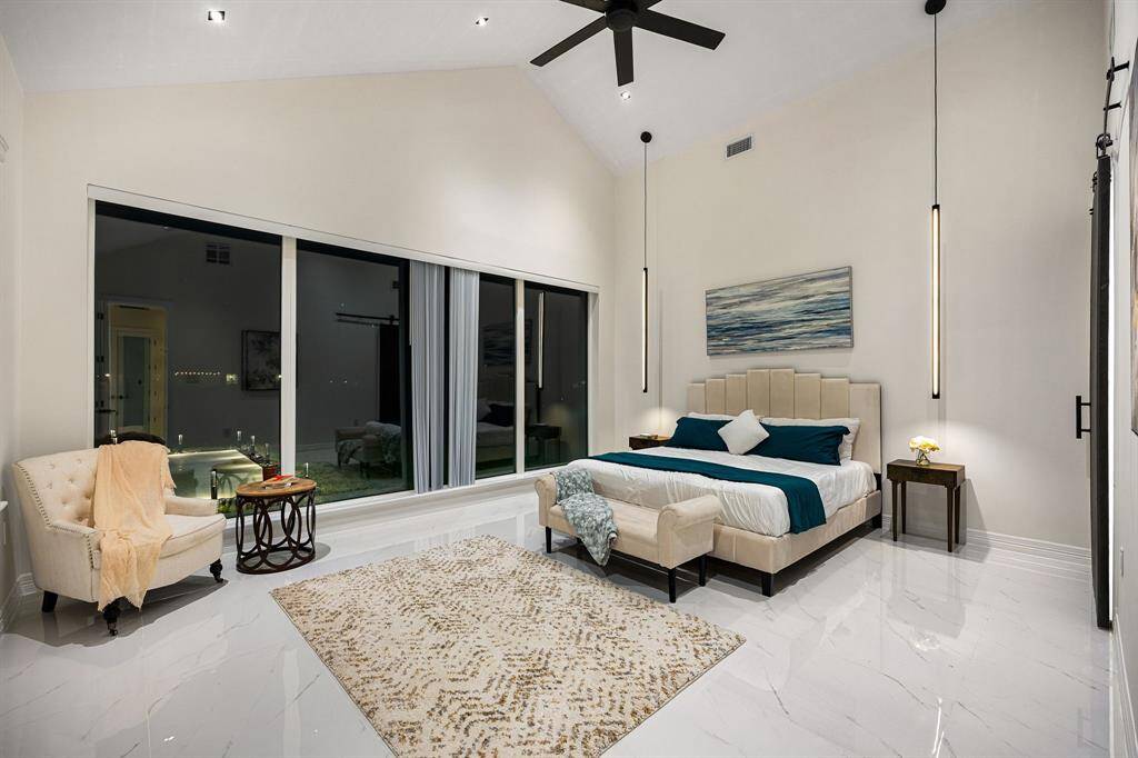 Indulge in the epitome of luxury in this exquisite primary suite, where dramatic vaulted ceilings and sophisticated pendant lighting set the tone for elegance. Floor-to-ceiling windows frame serene views of your private outdoor oasis, while gleaming marble-inspired floors and a soft, neutral color palette evoke a sense of calm sophistication. This is more than a bedroom—it’s your personal sanctuary.