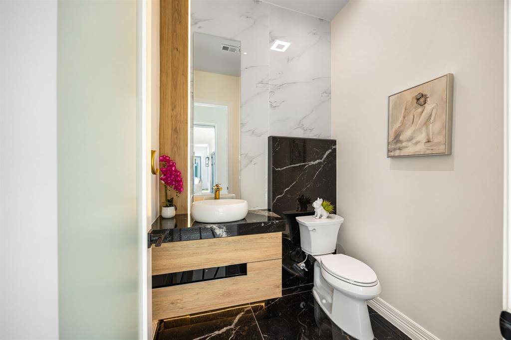 This chic half bath makes a bold statement with dramatic black marble flooring, warm wood finishes, and sleek gold accents. A vessel sink and backlit mirror add modern sophistication, while artistic touches elevate the overall ambiance—perfect for impressing guests with style and elegance.