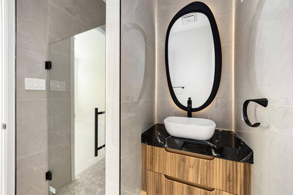 This stylish en suite bath features contemporary elegance with sleek gray tile walls, a bold black-framed mirror, and a striking vessel sink set atop a rich wood vanity with black marble countertop. A frameless glass shower and modern matte black fixtures complete the sophisticated, spa-like design.