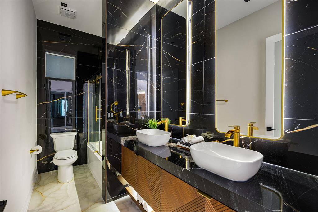 This striking secondary bathroom features bold black marble-style walls with gold veining, elevated by modern gold fixtures and dual vessel sinks. The large, illuminated mirrors and sleek wood vanity add a luxurious touch, while a glass-enclosed tub and shower combo offers both style and functionality.