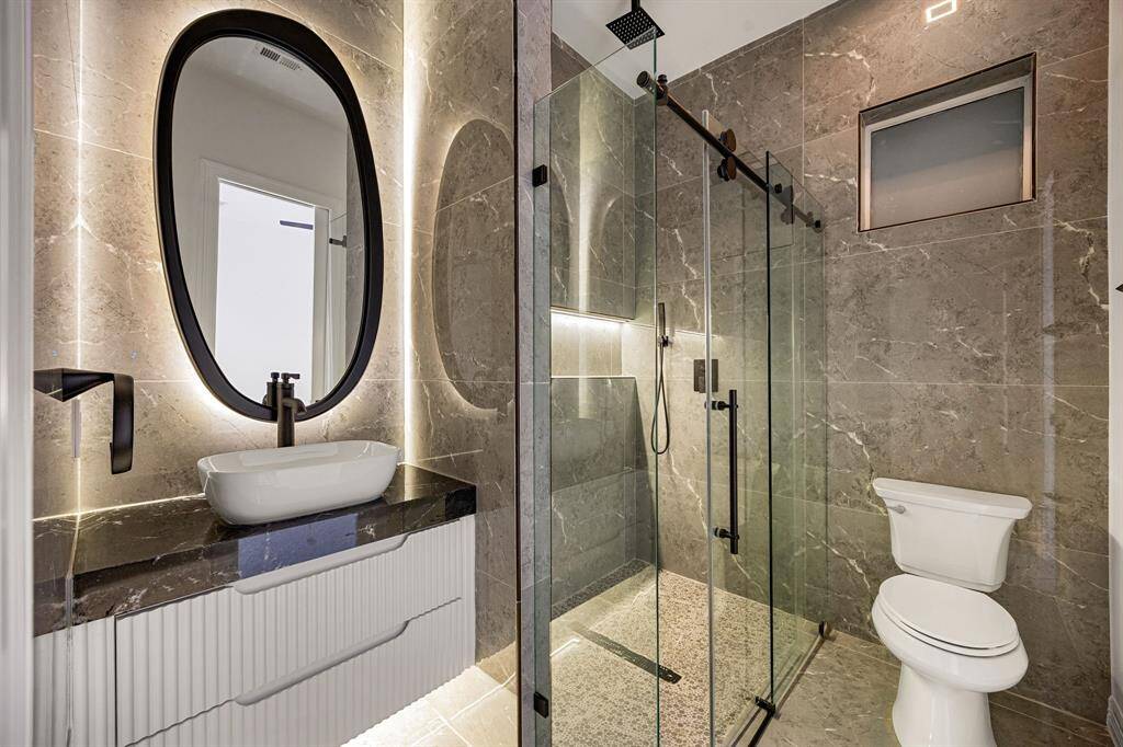 This stylish shared bathroom showcases a modern design with floor-to-ceiling stone tile, a sleek glass-enclosed shower with a rainfall showerhead, and a floating vanity topped with a vessel sink. Backlit mirrors and matte black fixtures add a touch of elegance and contemporary flair.