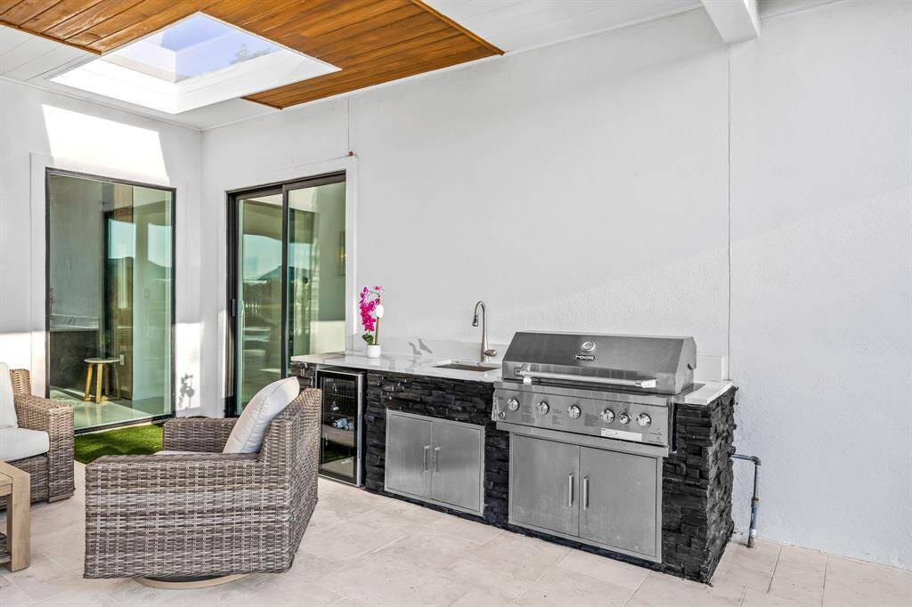 This covered outdoor kitchen is a chef’s dream, featuring a built-in stainless steel grill, sink, and mini fridge. A sleek skylight above brings in natural light, creating the perfect spot for cooking and entertaining in any season.