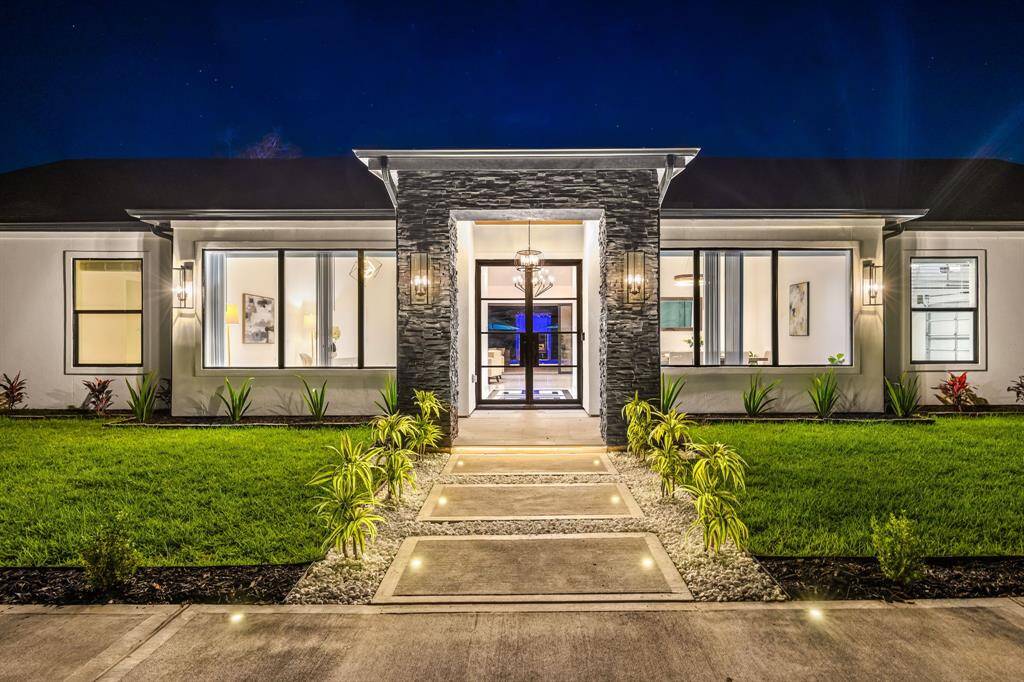 This stunning single-story residence showcases sleek architectural lines, expansive picture windows, and a grand stone-accented entryway illuminated by warm, ambient lighting. The manicured landscaping and illuminated walkway create an inviting path to the glass-paneled front door, offering a glimpse of the sophisticated interiors beyond. Designed for luxurious living and effortless entertaining, this home blends contemporary style with timeless comfort.