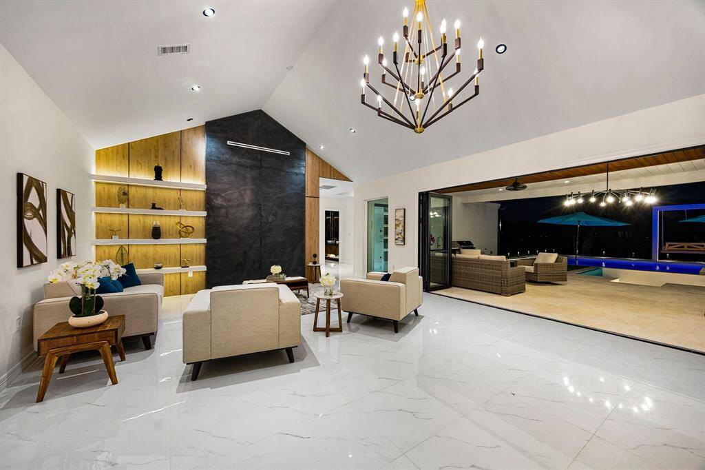 This striking living area impresses with its soaring vaulted ceiling, dramatic black stone accent wall, and custom built-in shelving with warm wood tones and integrated lighting. Large-format polished tile floors enhance the sense of space and luxury, while retractable glass walls blur the line between indoor and outdoor living, opening to a covered patio and an illuminated pool oasis beyond. A perfect blend of contemporary design and functional elegance.