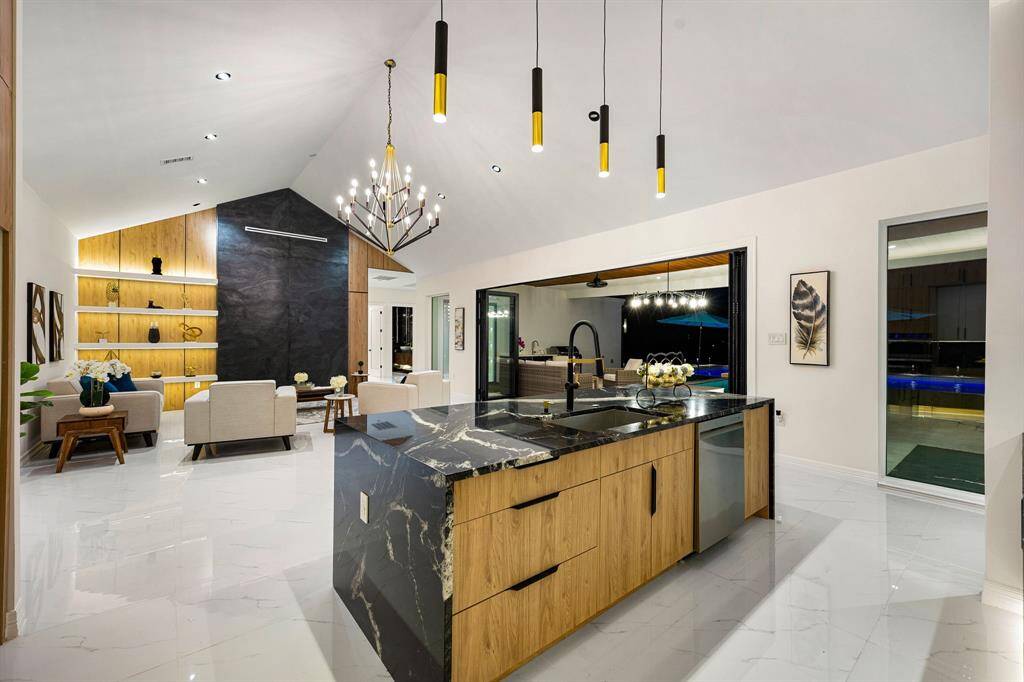 The kitchen features a bold waterfall island with luxurious black granite countertops and natural wood cabinetry, accented by contemporary pendant lighting. Large sliding glass doors seamlessly connect the indoor space to an inviting outdoor area, perfect for entertaining. Polished marble flooring and integrated lighting throughout enhance the bright, airy atmosphere, while a striking feature wall adds a sophisticated focal point to the living space.