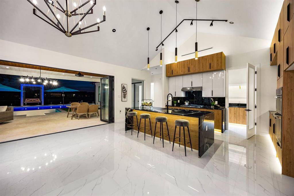 This breathtaking kitchen offers a seamless blend of modern luxury and functionality, highlighted by dramatic black granite countertops and a waterfall island. High-gloss cabinetry paired with warm wood creates a striking contrast, while pendant and track lighting add a sophisticated ambiance. Expansive sliding doors open to a resort-style outdoor space with pool views, enhancing the indoor-outdoor living experience. Gleaming marble floors and clean architectural lines complete the contemporary design.