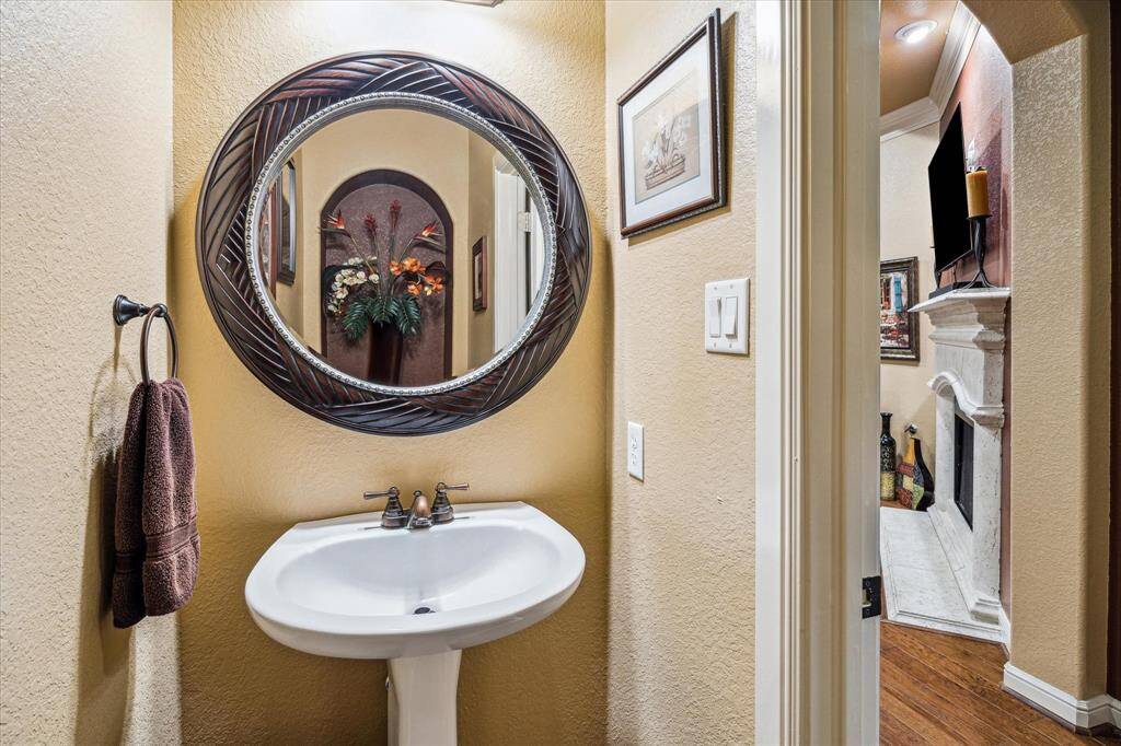 This charming powder room features a pedestal sink, warm neutral wall tones, and a decorative round mirror that serves as a stylish focal point. Bronze fixtures and tile flooring complete the space with a balance of elegance and functionality.