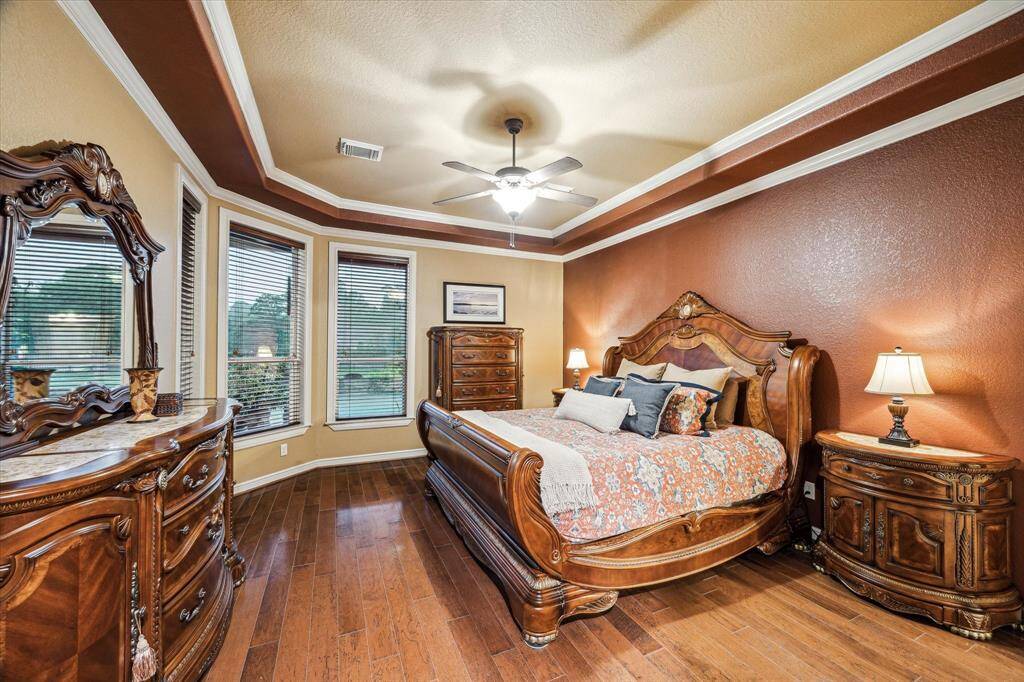 The serene primary bedroom suite features rich wood flooring, a tray ceiling with crown molding, and a striking accent wall. A wall of windows offers peaceful views of the backyard oasis, bringing in natural light and a sense of retreat.