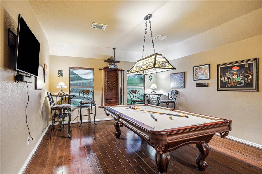 This spacious upstairs game room features rich wood flooring, soft neutral tones, and plenty of room for recreation and relaxation. Large windows bring in natural light, while recessed lighting and a custom pool table light add ambiance. Whether used for billiards, movie nights, or casual gatherings, this versatile space is ideal for entertaining family and friends.