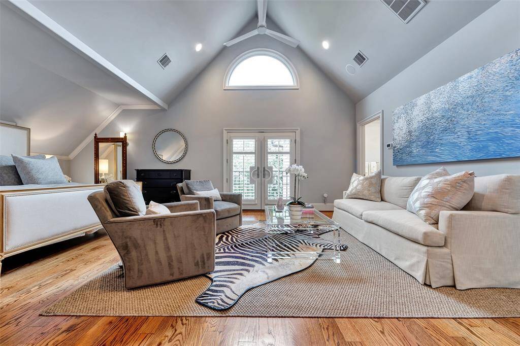 The spacious primary bedroom impresses with soaring vaulted ceilings, a large arched window, and French doors that flood the room with natural light. Elegant and serene, it offers a sitting area, hardwood floors, and refined architectural charm.
