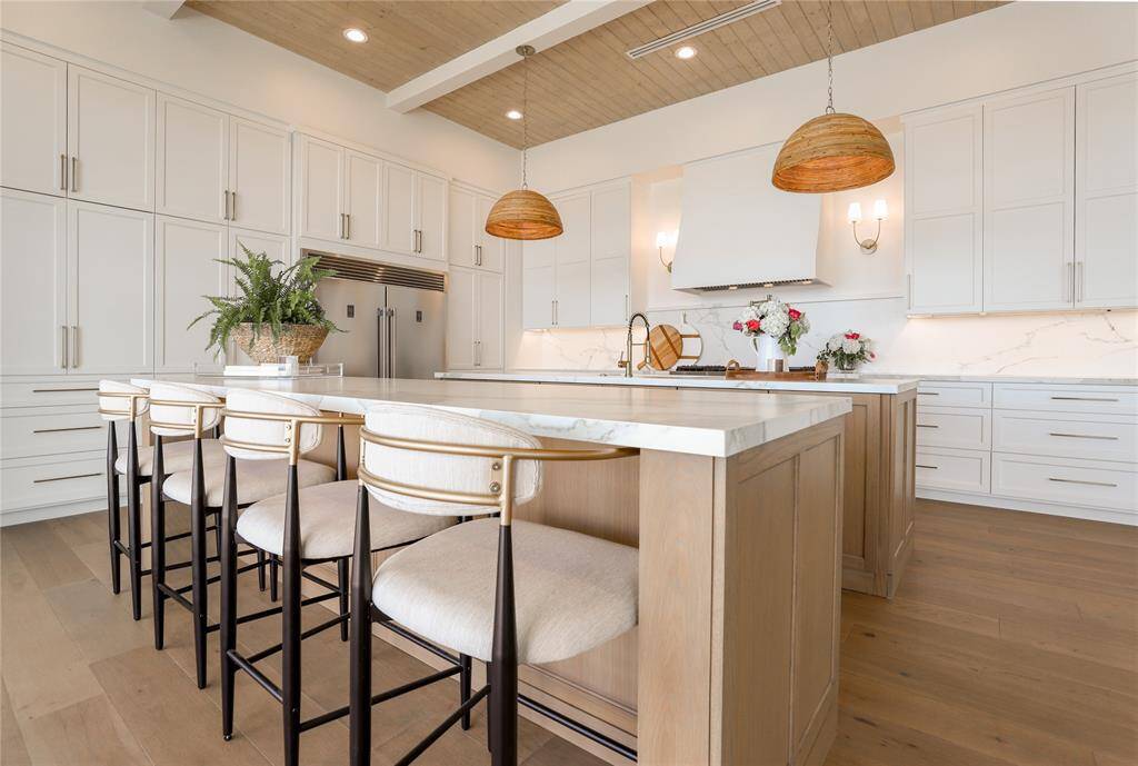 This modern kitchen features a spacious island with seating, sleek white cabinetry, and stainless steel appliances. The space is accented with warm wood tones, pendant lighting, and elegant Neolith countertops, creating a bright and inviting atmosphere.
