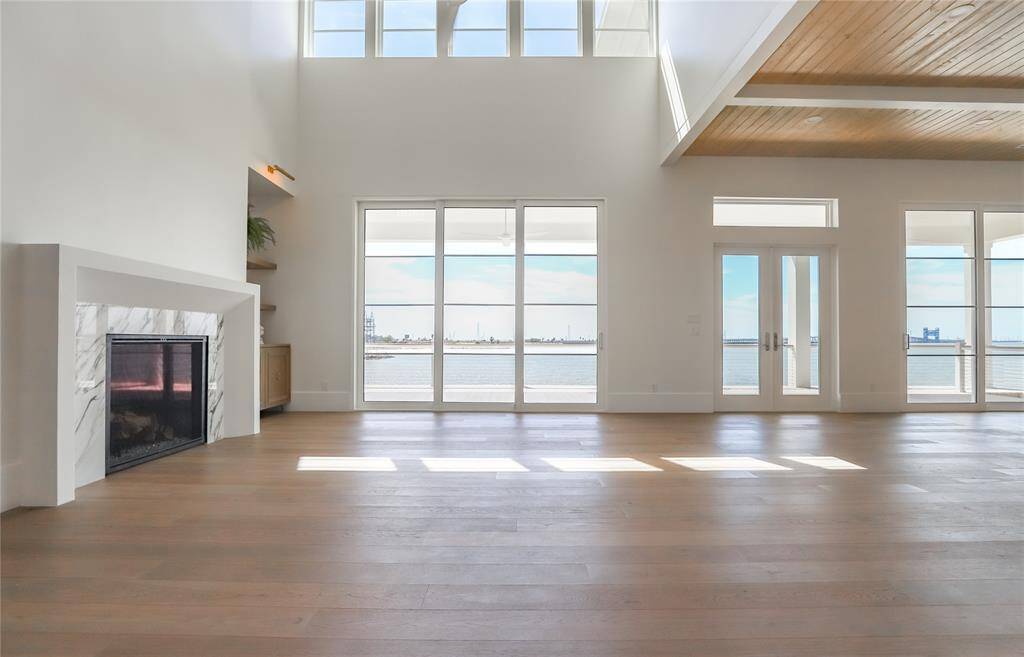 This spacious living area features high ceilings, abundant natural light from large windows, a cozy fireplace, and stunning water views, creating an inviting and airy atmosphere.