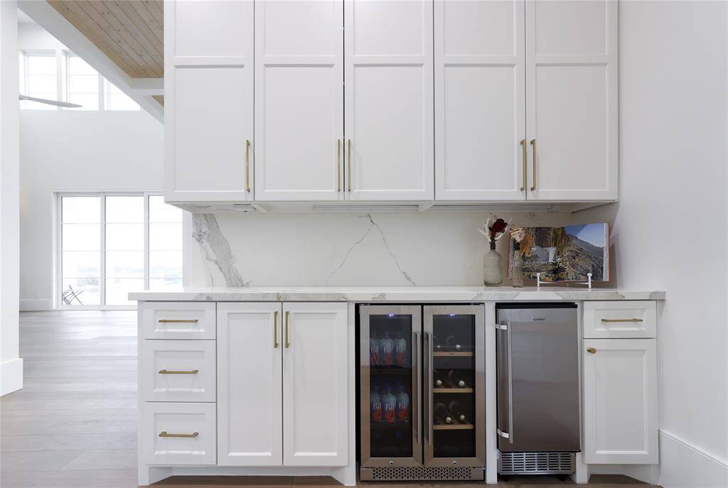 Discover a sleek modern dry bar featuring elegant Neolith countertops, a built-in beverage fridge, and an ice maker. The white cabinetry with gold handles enhances the sophisticated ambiance, complemented by ample natural light and an open layout.