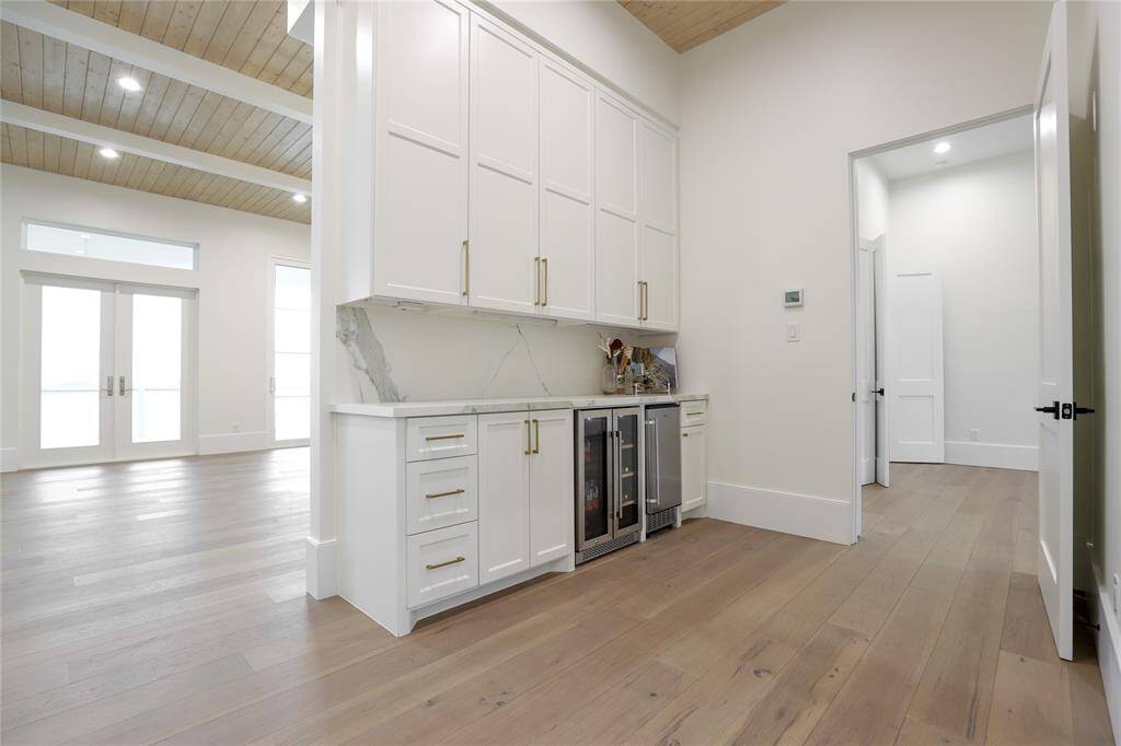 This modern home features an open space with high ceilings and recessed lighting. It includes a sleek beverage refrigerator and ice maker area with white cabinetry, and gold accents. The light wood flooring and large windows provide a bright, airy atmosphere.