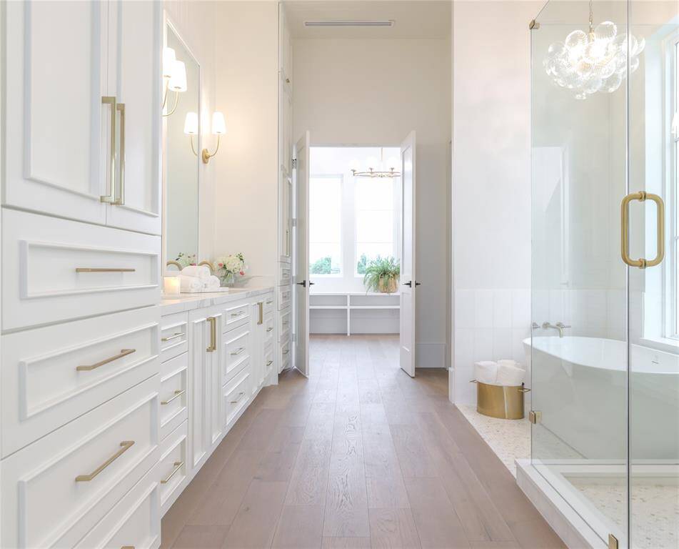This first level primary bathroom features a spacious, bright design with light wood flooring, ample white cabinetry, and elegant gold hardware. It includes a large glass-enclosed shower, a freestanding bathtub, and dual sinks. Natural light floods in through large windows, enhancing the airy feel.
