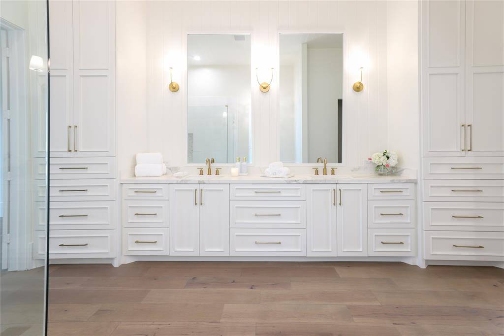 This elegant first level primary bathroom features a double vanity with sleek white cabinetry and gold hardware. It includes two large mirrors, stylish sconces, and ample storage. The neutral tones and contemporary design create a luxurious, spa-like atmosphere.
