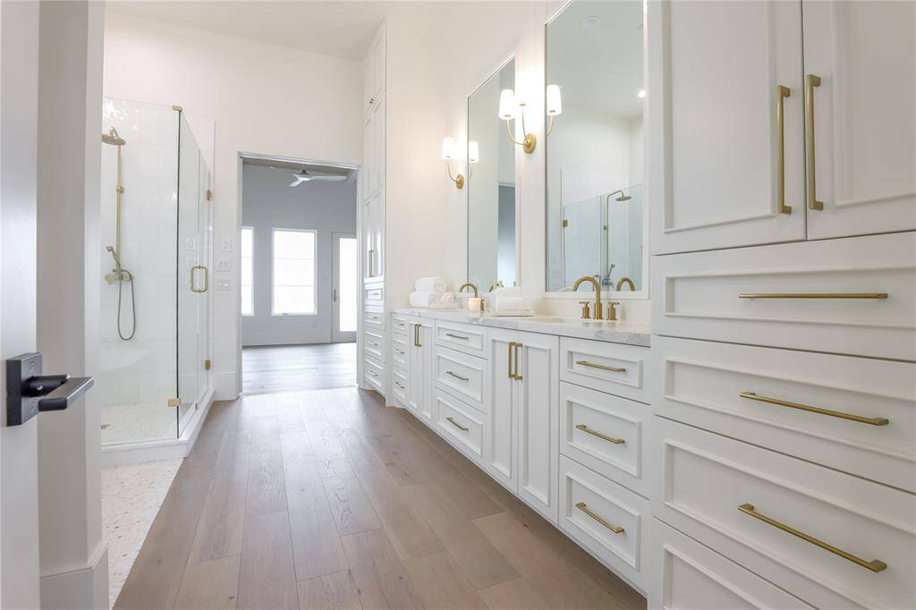 This photo showcases a spacious, modern bathroom with a double vanity featuring elegant gold fixtures and ample storage. The room includes a large glass-enclosed shower and opens into a bright bedroom with hardwood floors.