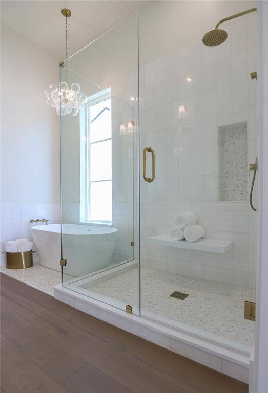 This first level primary bathroom features a modern design with a spacious glass-enclosed shower, a freestanding bathtub, elegant light fixtures, and wood flooring, creating a luxurious and relaxing space.