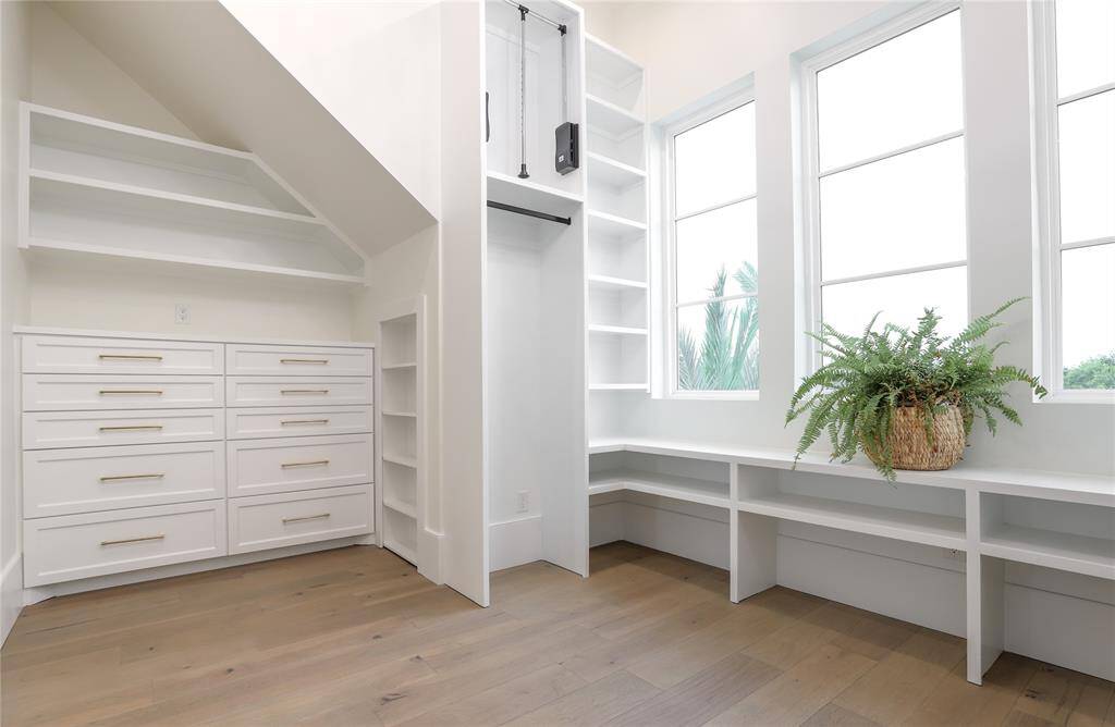 This bright, spacious first level primary bedroom closet features ample built-in storage with drawers and shelves, large windows for natural light, and a cozy nook with a stylish wooden floor.