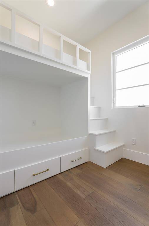 This photo showcases a bright, 2nd level bedroom featuring 2 built in bunk beds. The space features wood flooring, a white staircase leading to the loft, and storage drawers beneath. Ample natural light enters through a nearby window.
