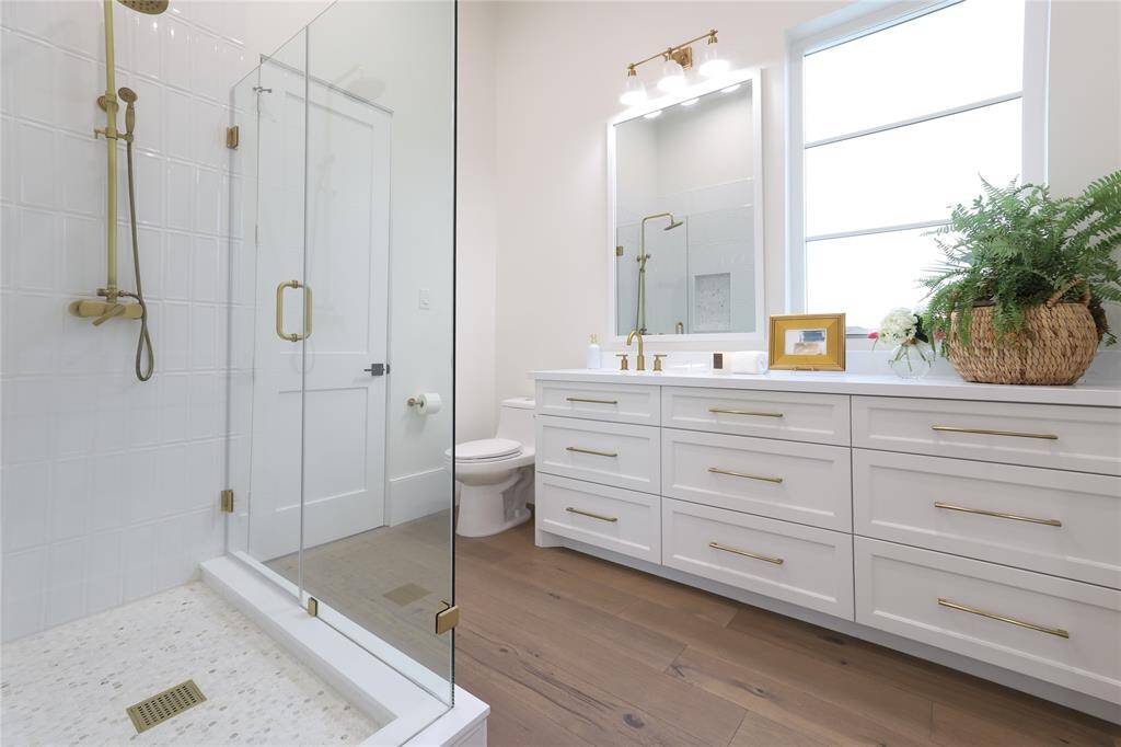This bathroom features a modern design with a spacious glass-enclosed shower, white cabinetry with gold hardware, and a large mirror. It has a clean, bright look with natural light from a window.  Located on first level with access to second bedroom and used as second floor bath for guests.