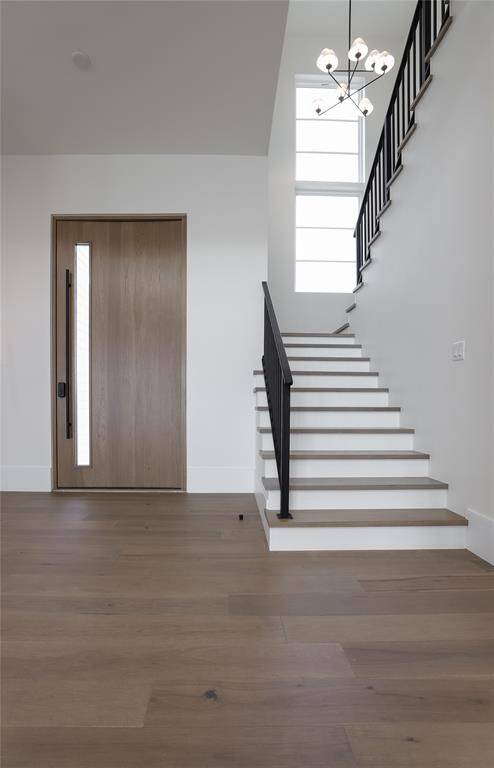 This modern entryway features a sleek wooden door with a vertical window, light wood flooring, and a stylish staircase with black railing. A contemporary chandelier adds elegance, and large windows provide ample natural light.