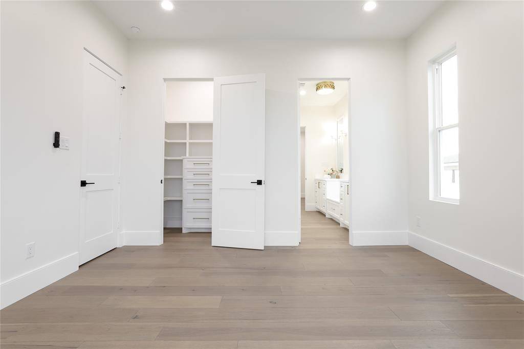 This photo shows a bright, modern second floor bedroom with light wood flooring and white walls. It features a walk-in closet with built-in shelves and drawers, and a view into a bathroom with dual sinks and ample lighting. Natural light enters through a window on the right.