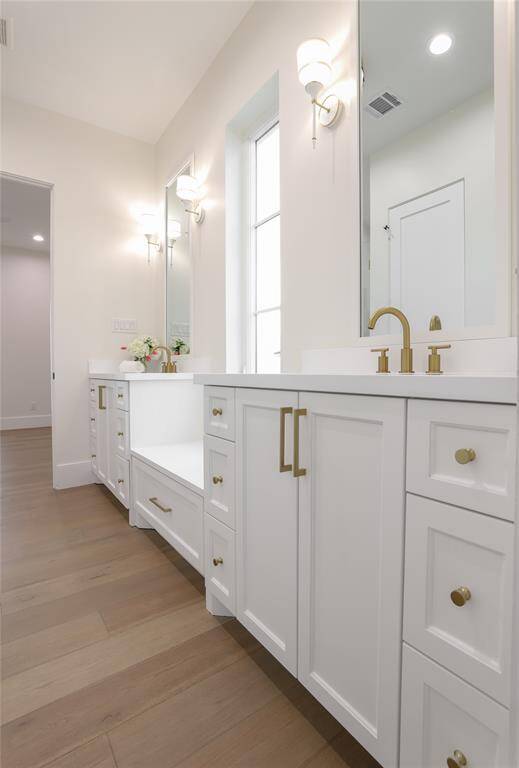 This bathroom features elegant white cabinetry with gold hardware, dual sinks, and large mirrors. The space is brightened by wall sconces and natural light from a window, complemented by light wood flooring for a modern, airy feel.  Jack and Jill bathroom shared with 2 bedrooms on 2nd floor.
