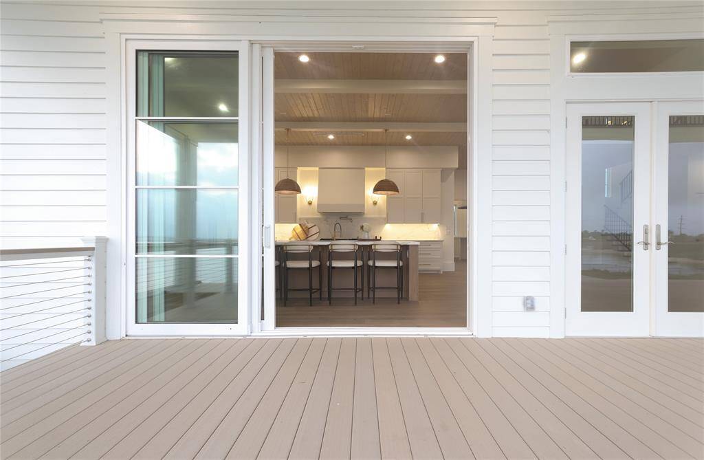 This photo showcases a spacious kitchen with an open layout, featuring modern cabinetry, pendant lighting, and a large island. It opens onto a light-colored deck through sliding glass doors, offering an inviting indoor-outdoor flow.