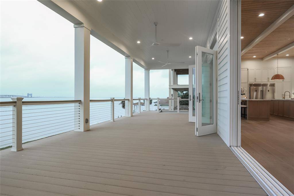 This photo showcases a spacious, covered deck with a stunning waterfront view. The deck features ceiling fans, recessed lighting, and a seamless transition through large glass doors to a modern kitchen with high ceilings and elegant finishes. Perfect for enjoying outdoor living and entertaining.