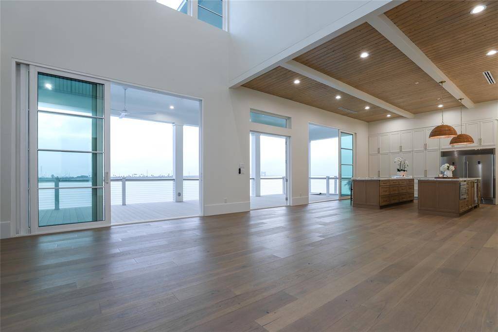 This modern open-concept living area features hardwood floors, high ceilings, and abundant natural light. It includes a spacious kitchen with a wooden ceiling, stylish pendant lighting, and large windows leading to a deck with water views.