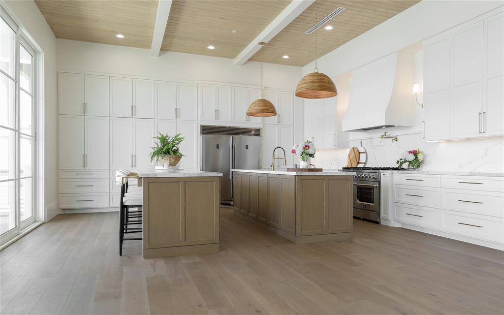 This kitchen features a spacious, open layout with modern white cabinetry, a large island with seating, stainless steel appliances, and elegant pendant lighting. The wood flooring and ceiling beams add warmth, while ample natural light streams in through large windows.