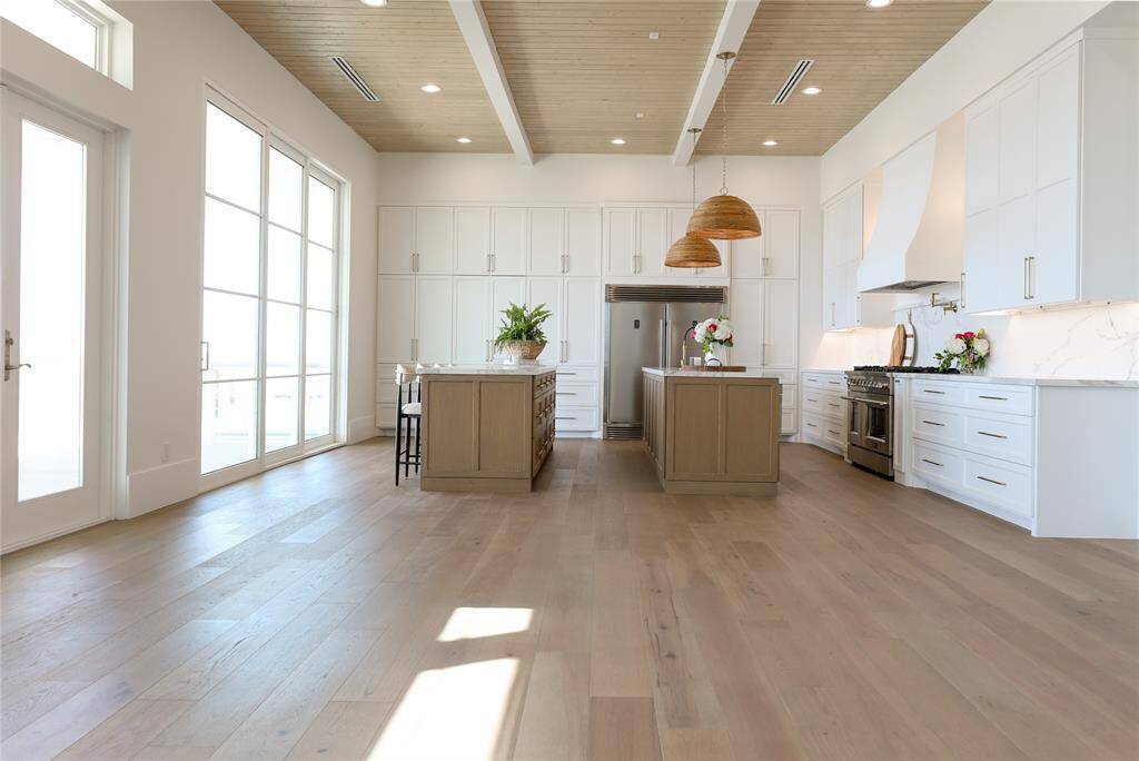 This photo showcases a spacious, modern kitchen with high ceilings and abundant natural light. It features sleek white cabinetry, a large central island with wooden accents, also features second island as breakfast bar for seating, Neolith countertops, stainless steel appliances, and a stylish range hood. The light wood flooring and ample windows create an inviting, airy atmosphere.