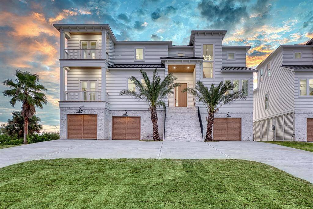 This modern, multi-story waterfront home with 7 bedrooms 5 baths features a sleek design with large windows, multiple balconies, and a spacious driveway leading to three garage doors. Accented with palm trees, its elevated entrance offers a grand appeal, set against a vibrant sunset backdrop.