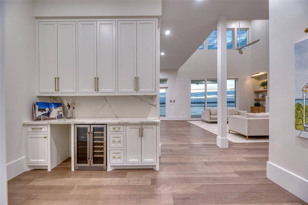 This modern, open-concept living space features elegant white cabinetry with a built-in beverage cooler, rich hardwood flooring, and expansive windows offering stunning water views.