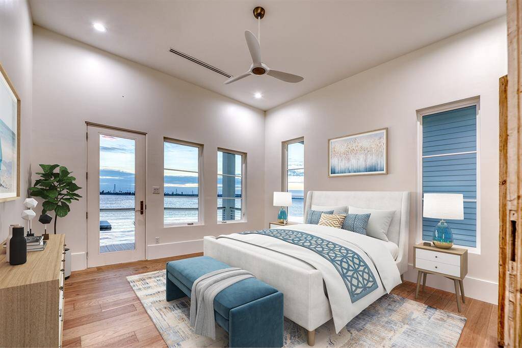 This serene first level primary bedroom offers stunning waterfront views through large windows and a glass door leading to a covered patio. It features warm wood flooring, a cozy bed with blue accents, and modern decor, creating a tranquil retreat.  Virtually staged.
