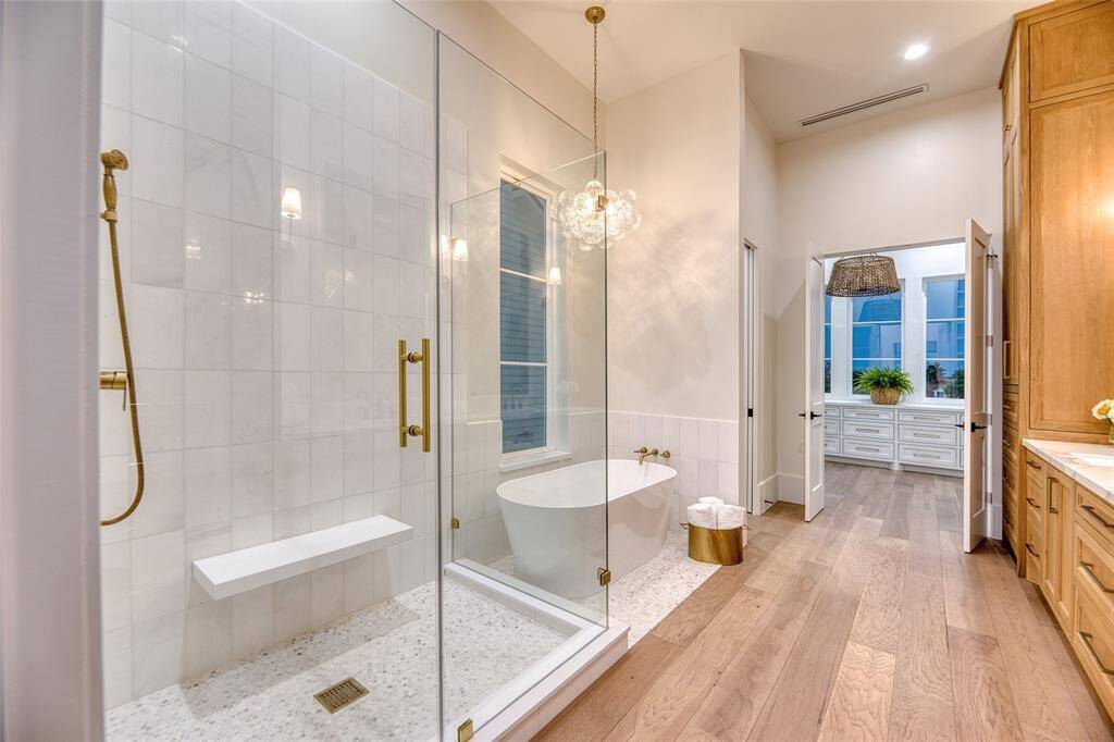 This spacious first level primary bathroom features a modern glass-enclosed marble shower with gold fixtures and a built-in bench, a stylish free-standing soaking tub, and elegant wood flooring. It opens into a bright room with large windows and ample storage, creating a luxurious and inviting atmosphere.