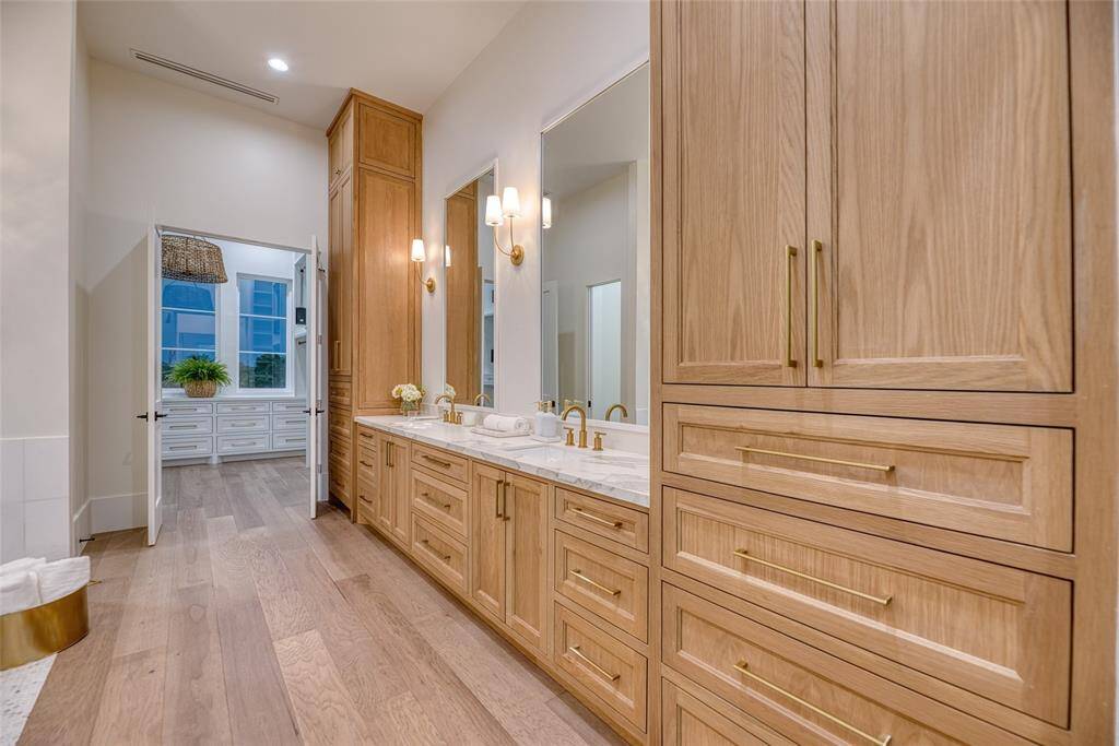 This photo showcases a spacious, elegant first level primary bathroom with light wood cabinetry, dual sinks, and gold fixtures. The room is well-lit with modern sconces and features a large, open doorway leading to a large walk-in closet. The hardwood flooring adds warmth to the space.
