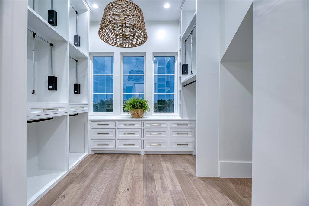 This photo showcases a spacious, modern first level primary bedroom walk-in closet with ample built-in storage, including drawers and hanging space. It features large windows for natural light, a wooden floor, and a stylish woven pendant light.