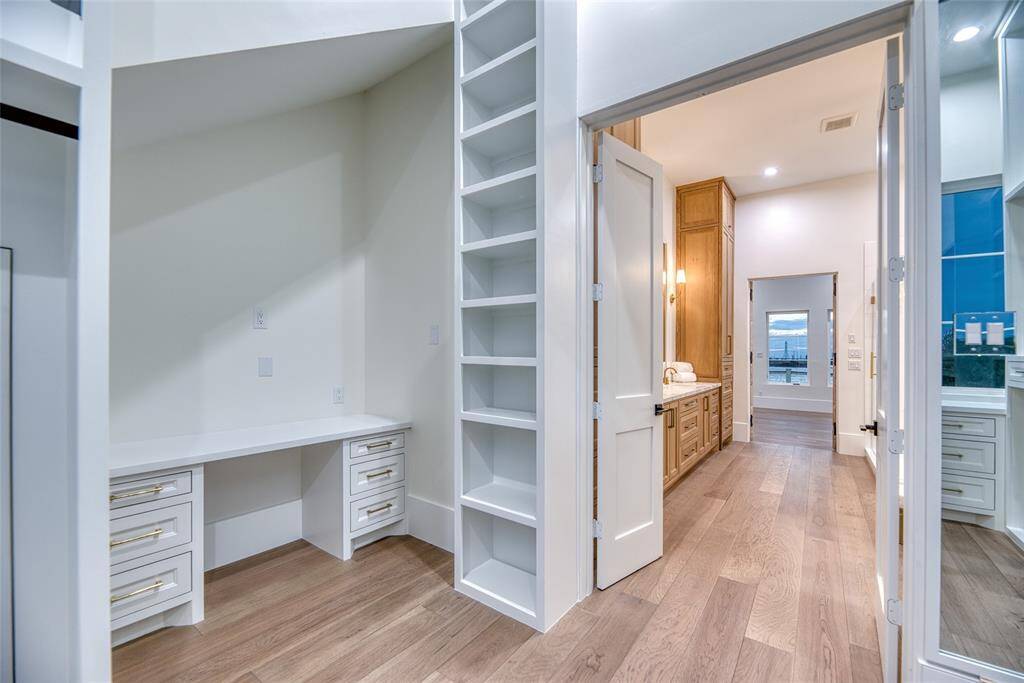 This photo showcases a bright, modern first level walk in primary bedroom walk in closet featuring a built-in vanity with storage drawers, open shelving, and hardwood floors. It leads to a hallway with a wooden cabinet and elegant lighting, creating a functional and stylish area.