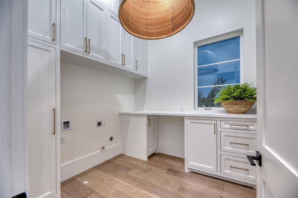 This photo shows a bright and modern laundry room with white cabinetry, ample storage, and a wooden floor. There's a large window for natural light and a stylish overhead light fixture. The room is clean and functional, ideal for laundry tasks.