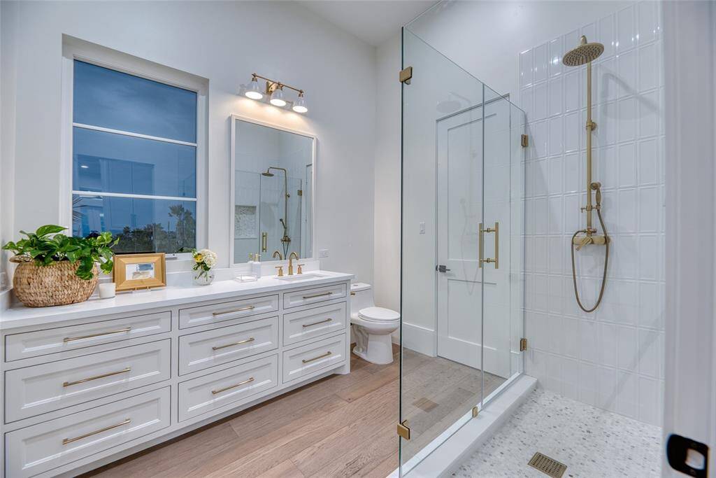 This first level bathroom features a spacious, modern design with a large glass-enclosed shower, elegant brass fixtures, and a sink with  vanity and ample storage. A large window provides natural light, and warm wood flooring.