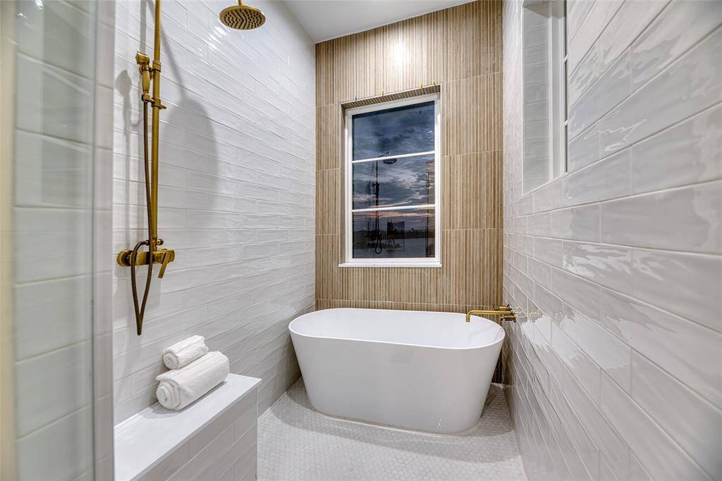 This modern second level bathroom features a sleek freestanding tub and a stylish shower with brass fixtures. The walls are lined with glossy white tiles, and a large window provides natural light, enhancing the spa-like atmosphere.