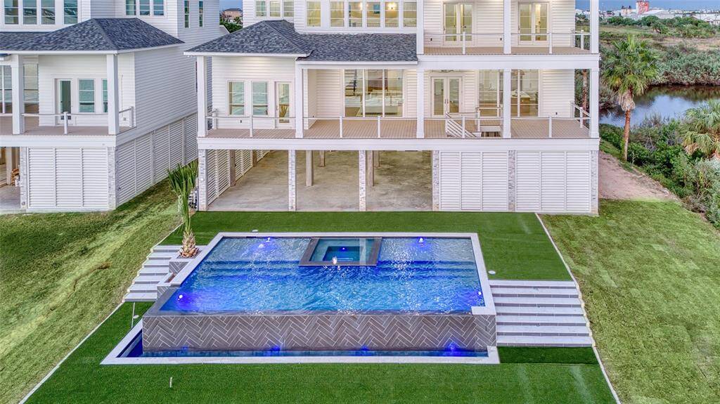 This modern, coastal waterfront home features a spacious backyard with a stunning infinity pool and hot tub, gas plug for firepit surrounded by lush landscaping. The property offers expansive outdoor living spaces and is designed to maximize views and natural light.