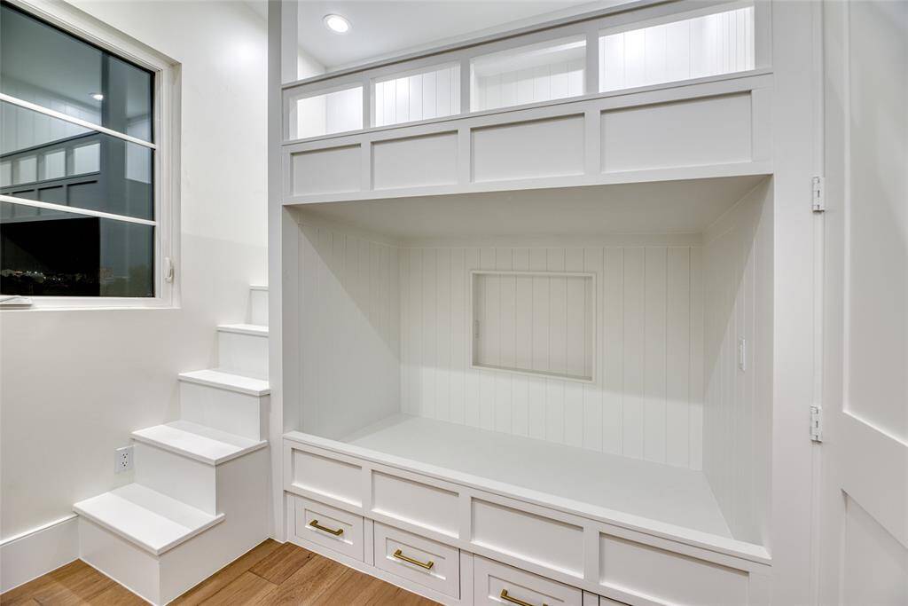 This cozy nook features built-in bunk beds with storage drawers beneath, a small staircase, and a window offering natural light. The white paneled walls and ceiling create a bright, inviting atmosphere. Perfect for a child's room or a playful guest space.