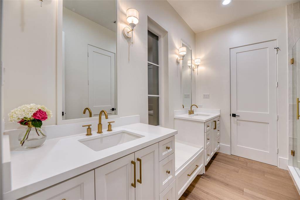 This second floor bathroom features a modern, elegant design with dual white vanities, gold fixtures, and ample mirror space. The wood-look flooring adds warmth, while soft lighting creates a cozy ambiance.