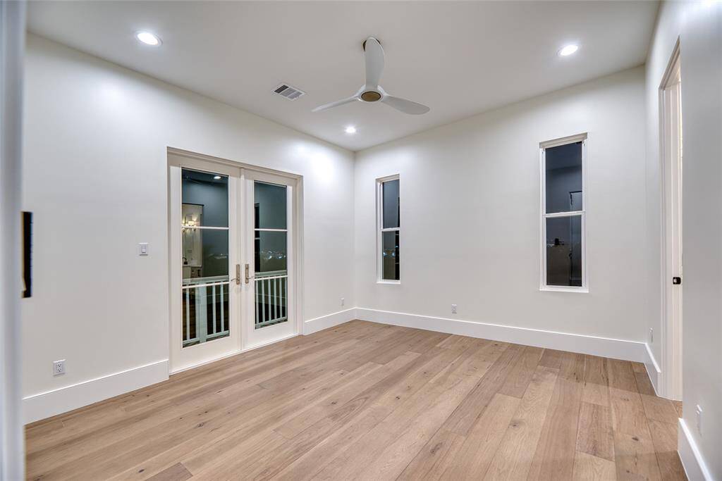 This second floor bedroom room features modern hardwood flooring, recessed lighting, and a ceiling fan. It has French doors leading to a small balcony and two tall windows that provide natural light. The neutral wall color complements the bright, open space.