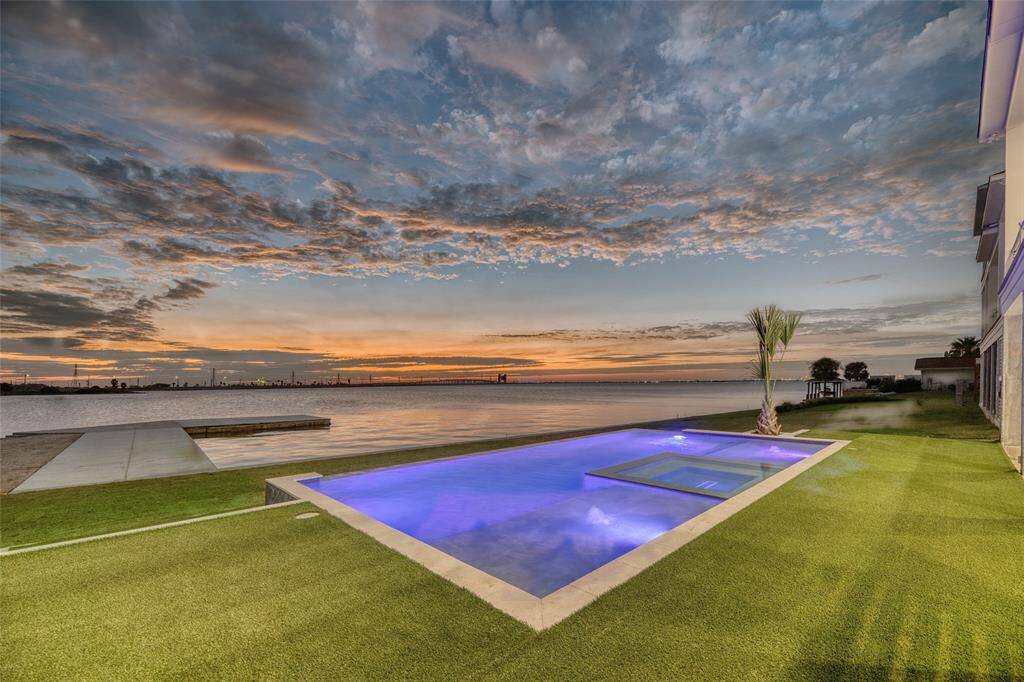 This stunning waterfront property features a modern infinity pool with ambient lighting, lush green lawn, and expansive views of a serene body of water, perfect for enjoying picturesque sunsets.
